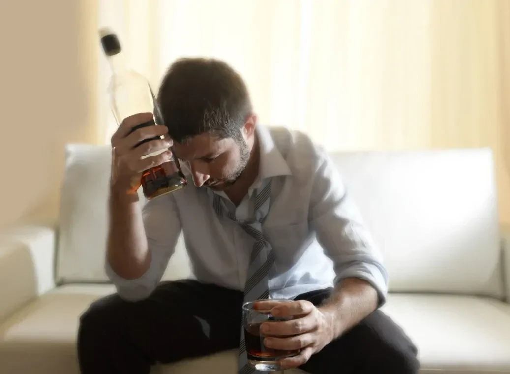 Mannelijke alcoholist zittend met fles drank tegen hoofd en een glas alcohol in zijn andere hand.