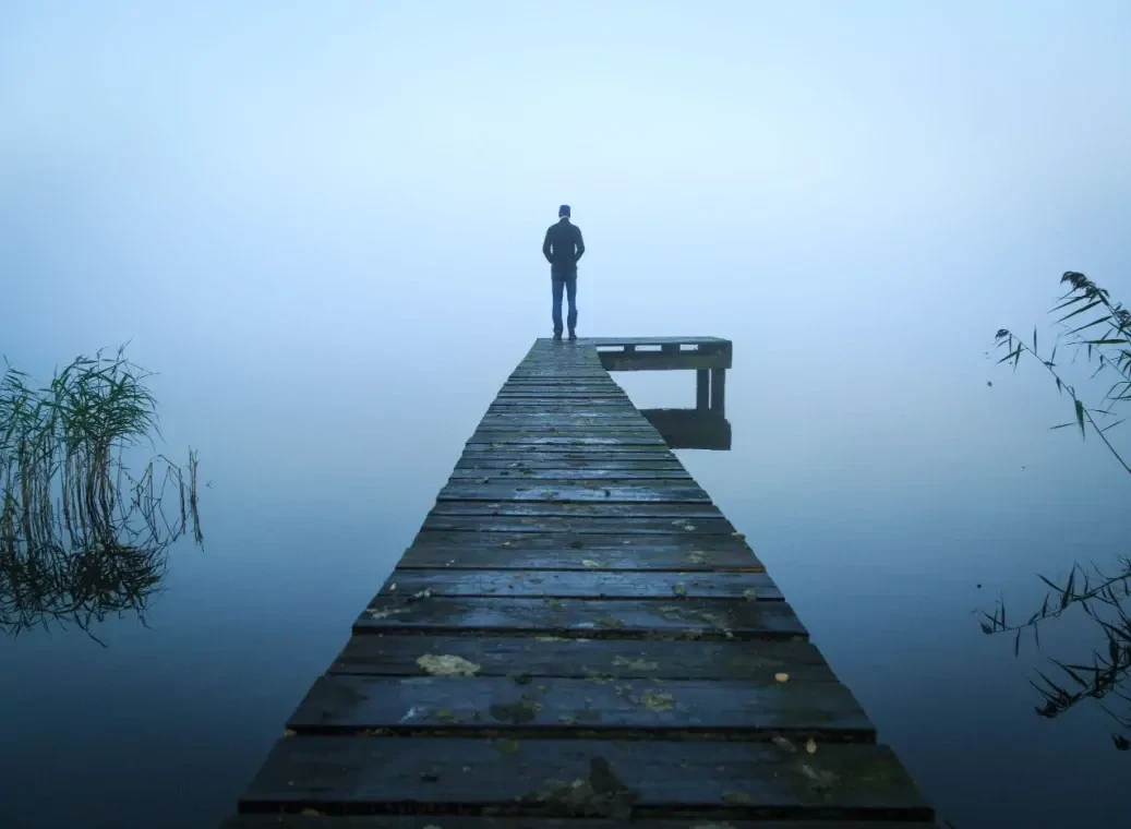 man-op-pier Een man staat in zijn eentje op een houten aanlegsteiger en kijkt uit over een mistig meer.