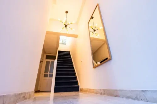 Hal met witte muren, een plafondlamp, een spiegel aan de muur en een donkere trap die naar boven leidt.
