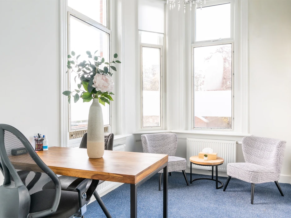 Behandelkamer met bureaustoel, houten bureau en twee fauteuils tegen hoge ramen en witte muren.