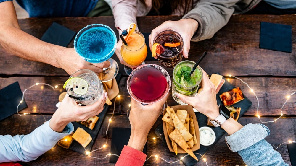 Groep jongeren die aan een picknicktafel borrelt, hapjes deelt en met cocktails proost, illustratie bij onderzoek naar alcoholgebruik onder jongeren.