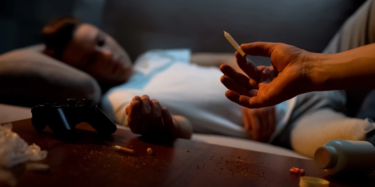Hand met joint bij persoon op bank met softdrugs en rommel op tafel.