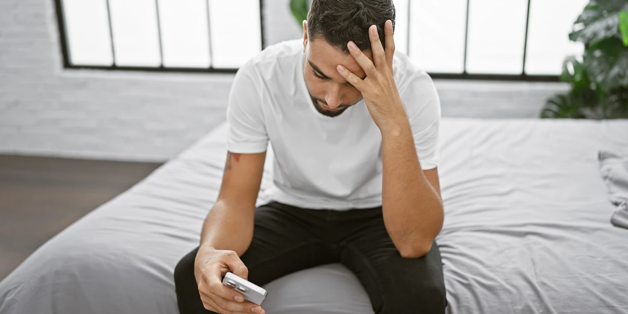 Man met ADD kijkt bezorgd naar telefoon terwijl hij op bed zit.