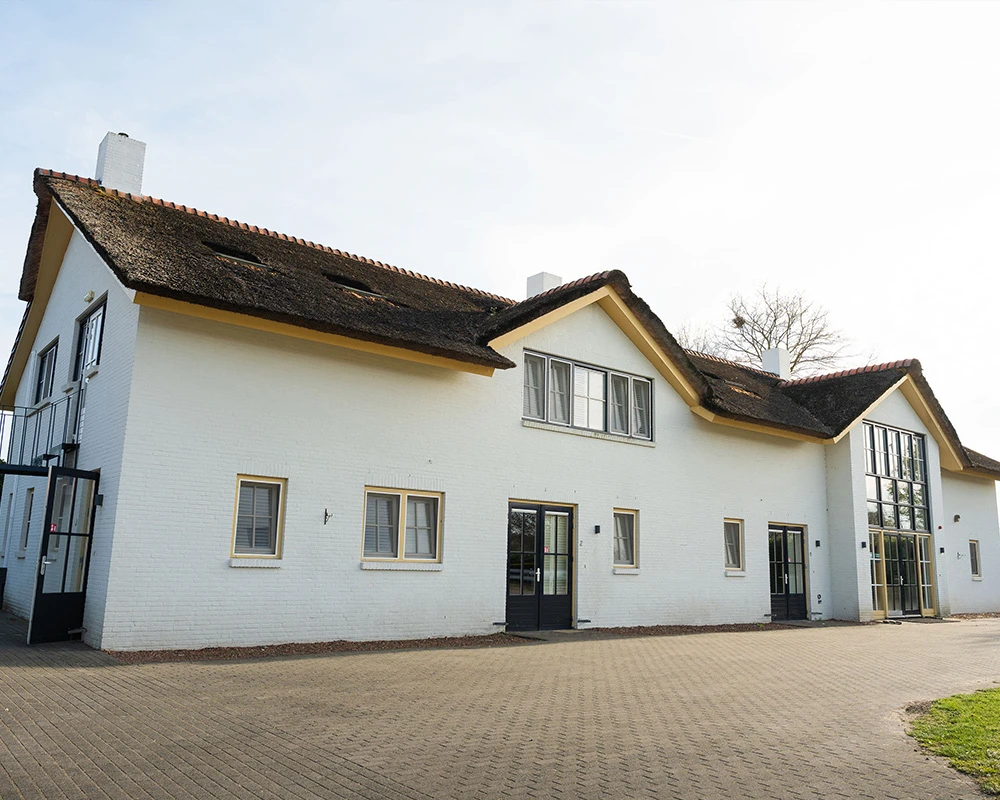 Vooraanzicht van afkickkliniek Meerlo, wit gebouw met rieten dak en grote ramen.