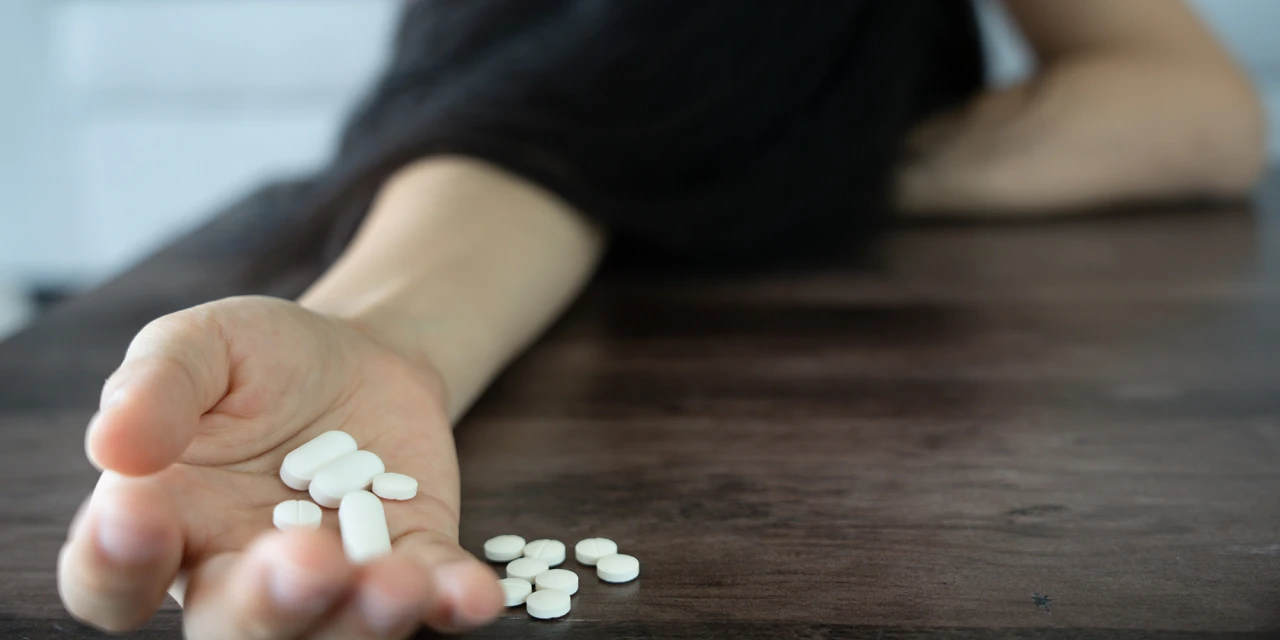Hand met pillen op tafel terwijl persoon uitgeput ligt door medicijnverslaving.
