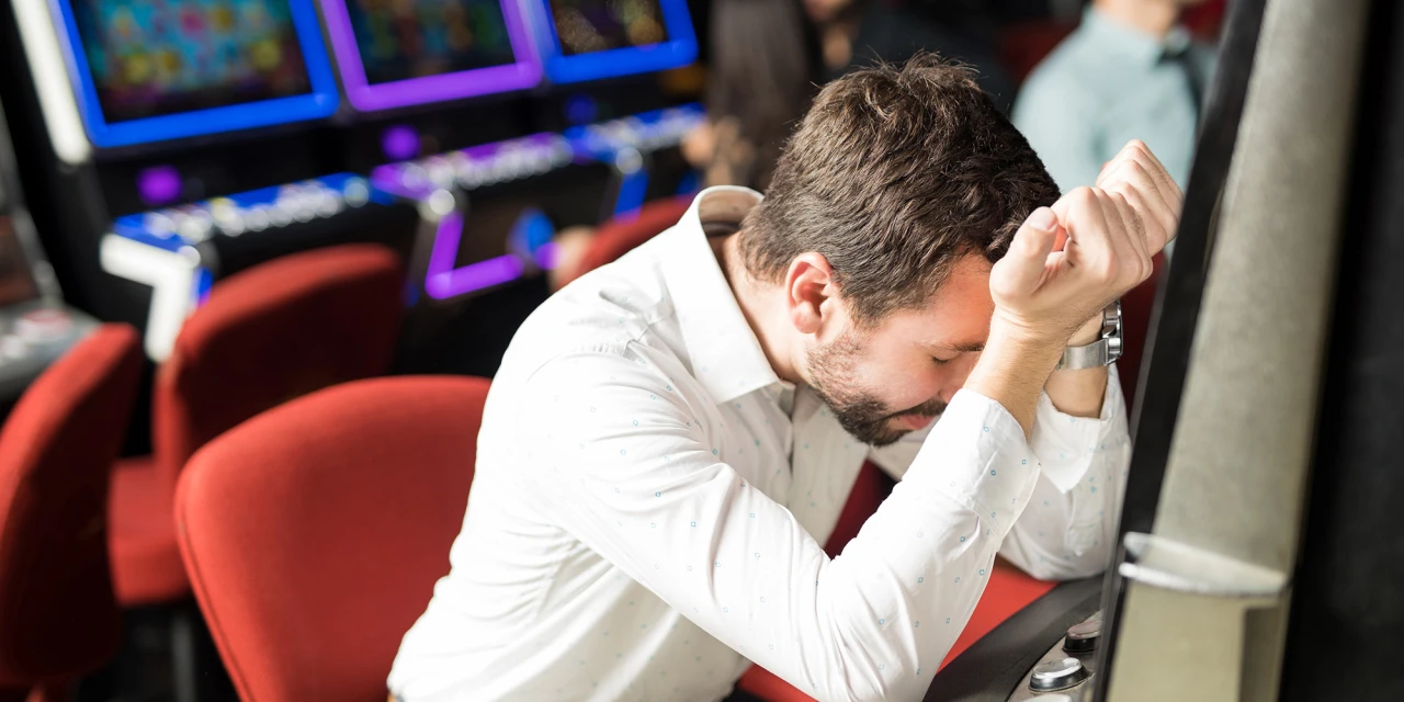 Man in casino leunt met hoofd op armen voor gokkast: afkickverschijnselen gokken zichtbaar.