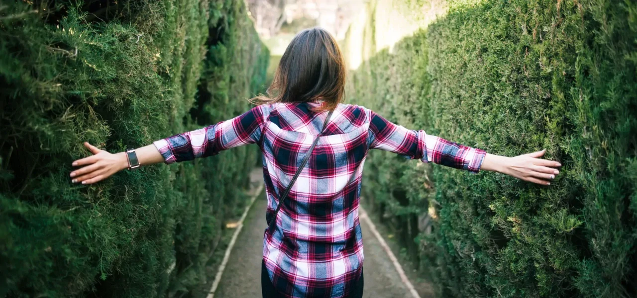 Achteraanzicht van vrouw met geruiten blouse loopt met haar armen gespreid door tussen twee groene heggen.