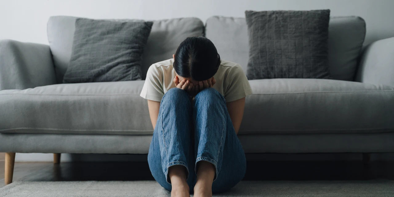 Vrouw zit met de handen voor haar hoofd verdrietig voor een bank op de grond. 