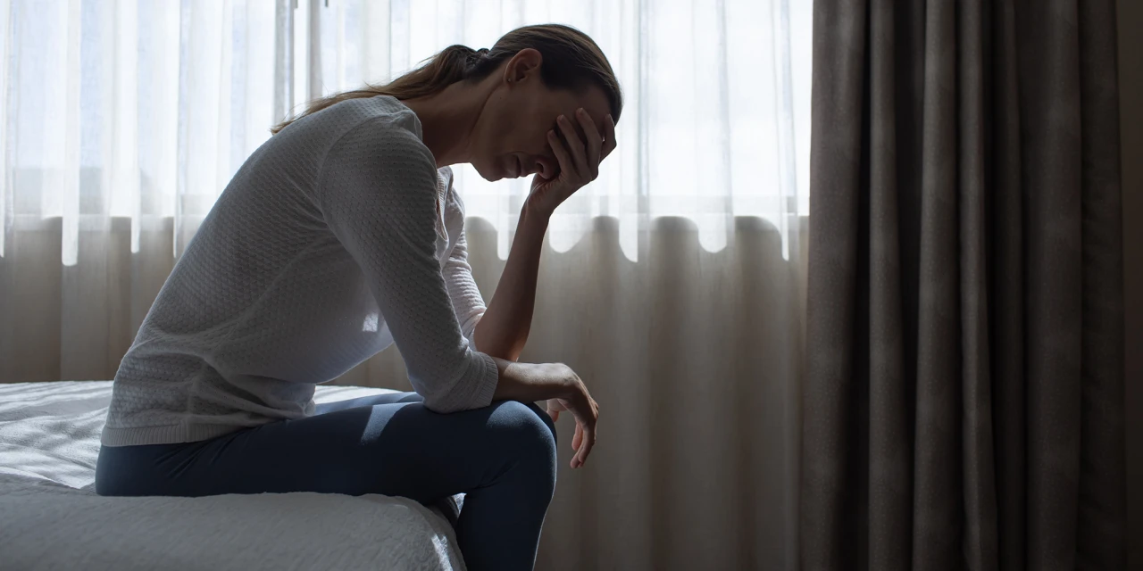 Vrouw zit op bed met hand op hoofd, zichtbaar vermoeid en somber.