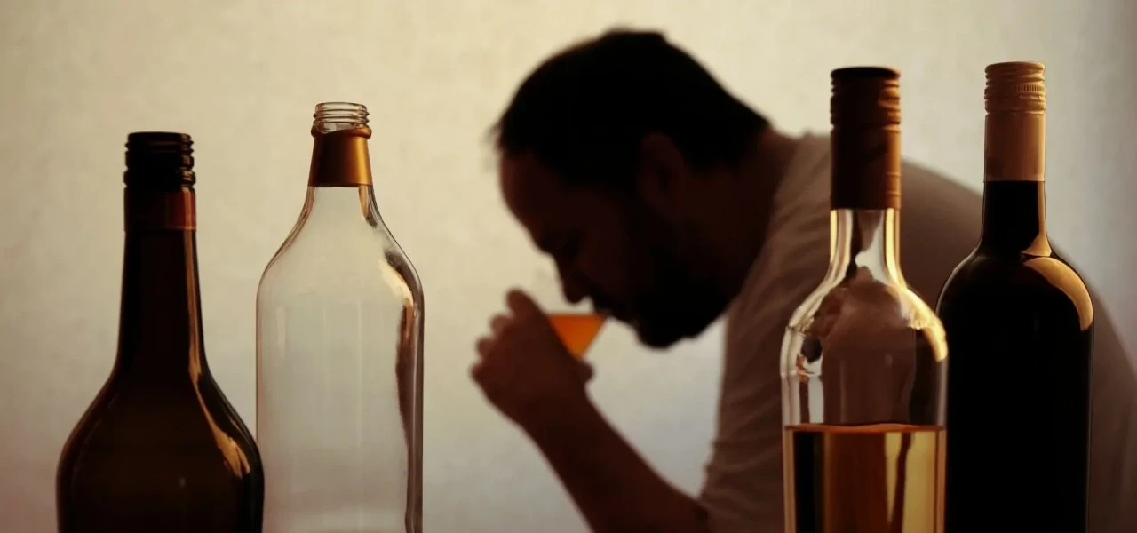 drinkende-man-met-alcoholabusus Man met alcoholabusus drinkt uit glas en heeft verschillende flessen drank voor hem staan.