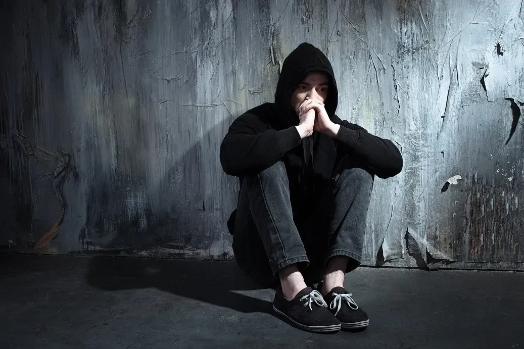 Depressieve-man-zittend Man zittend tegen een verweerde muur in een hoodie, met zijn handen gevouwen voor het gezicht.