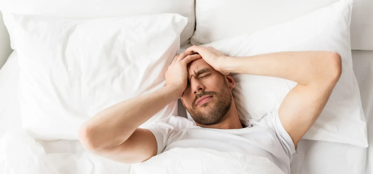 man-heeft-last-van-gevolgen-alcohol Man heeft last van de gevolgen van alcohol drinken en ligt in bed met handen op het hoofd.